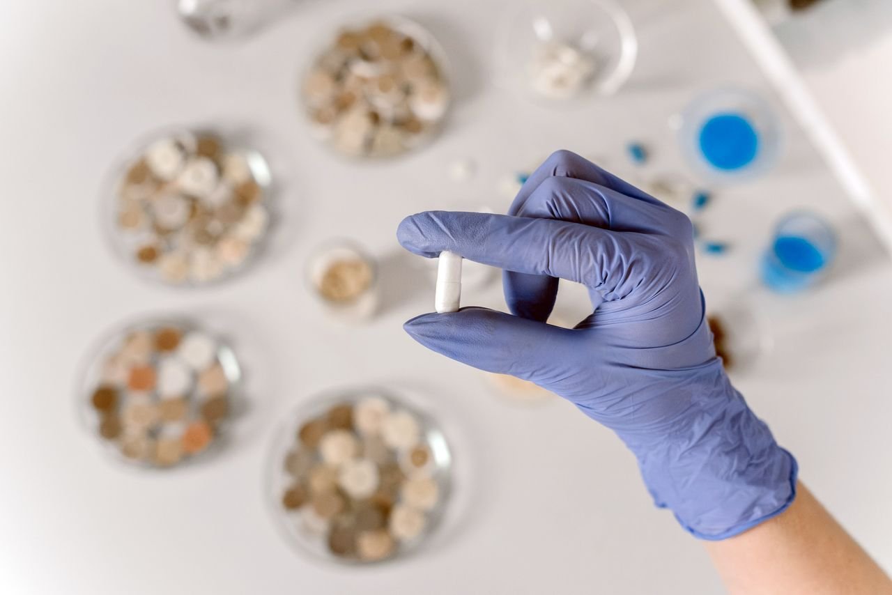 Close-up of a gloved hand holding a white capsule in a lab environment.